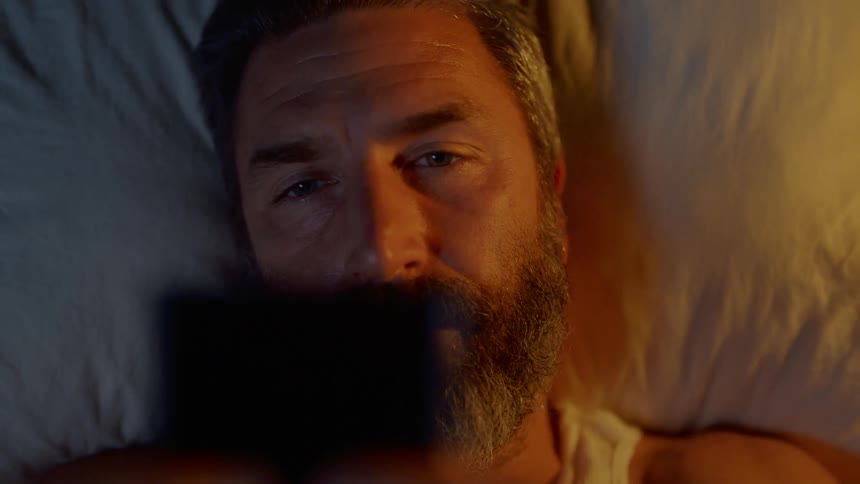 A man lays in bed and picks up a small card to look at it