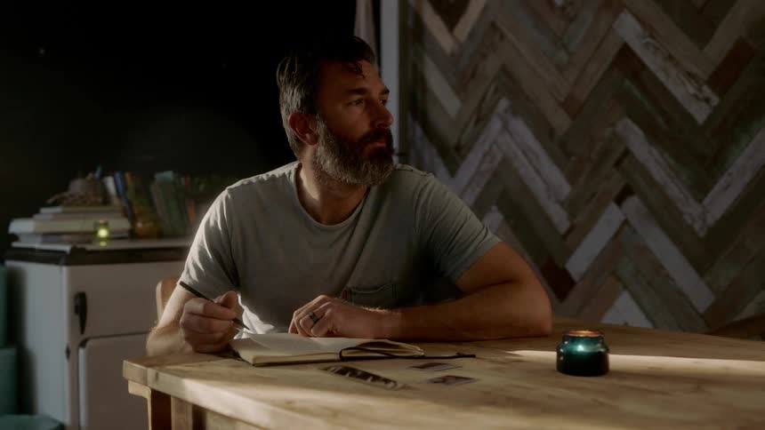 A man sits in a dark room writing in a notebook at a table