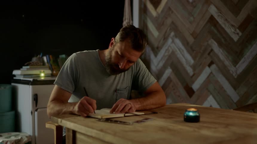 Man sits at wooden table near flickering candle writing