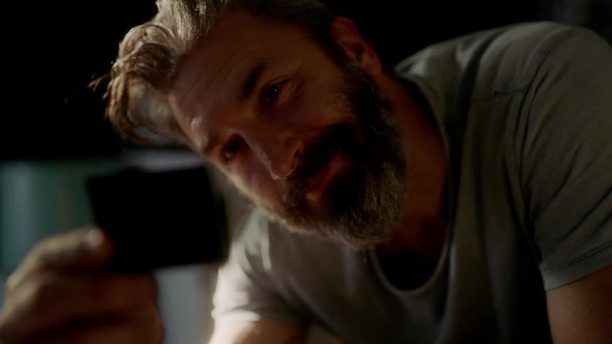 Male sits in a chair leans forward and tilts his head while smiling and looking at a picture
