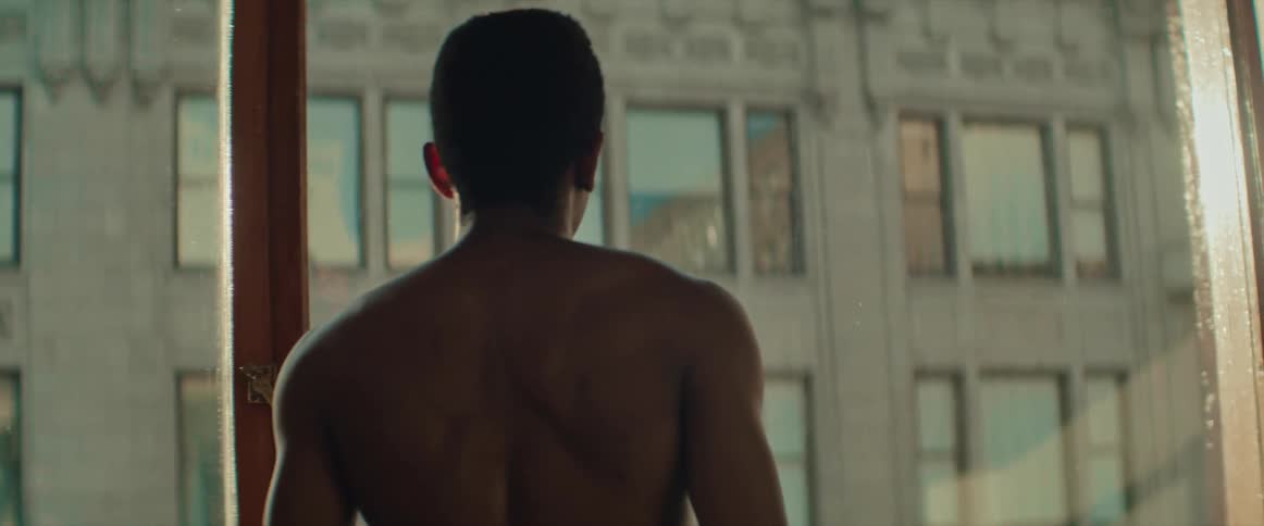 A shirtless man stands at a large window looking out and the windows of the opposite building reflect the sky and surroundings