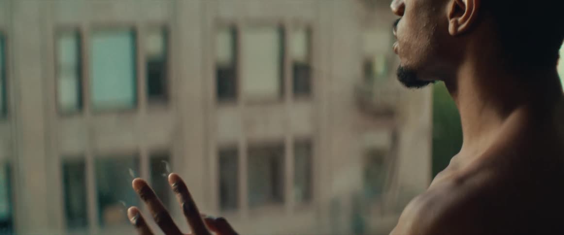 Shirtless black man touching the windowpane looks out the window in the apartment
