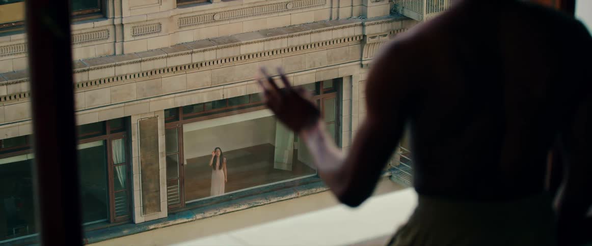 A shirtless man waves from his window to a woman in another window of an apartment building across the street