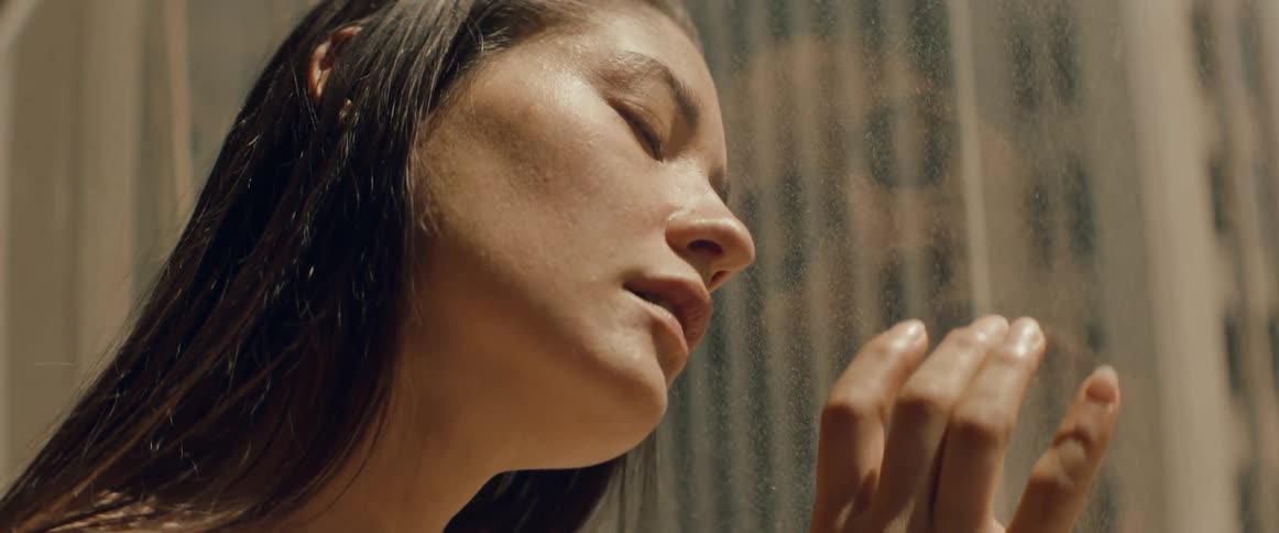 A woman rests her head against of the large window of her apartment and runs her hand down the glass