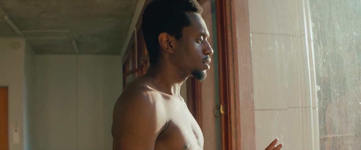 Shirtless man looks out the window at day