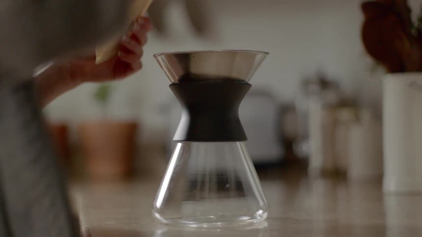 A person placing a coffee filter in the top of a glass coffee pot