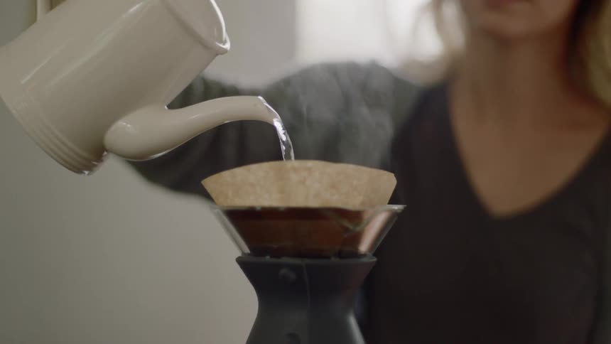 A woman adds hot water from a ceramic kettle to a pour over coffee maker at home