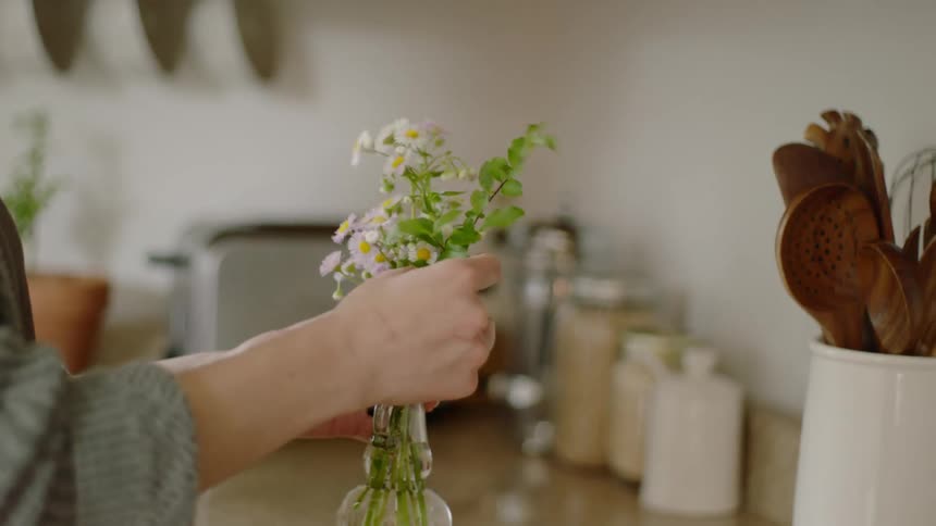 A woman places flowers in a small glass vase on the kitchen counter