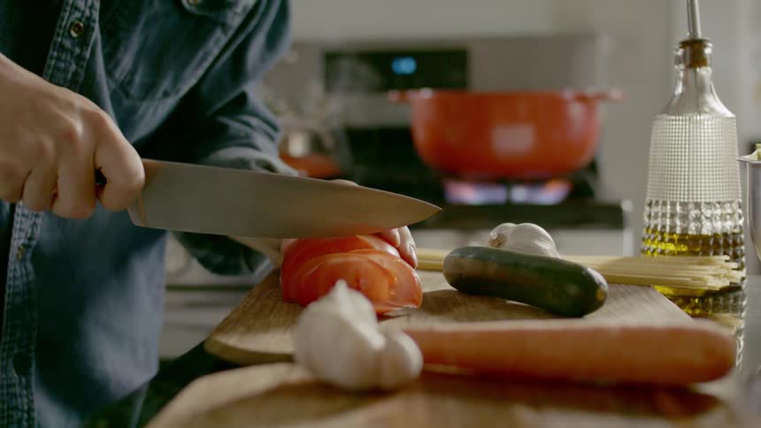 A person with a knife chopping a tomato with a pot on the stove in the background