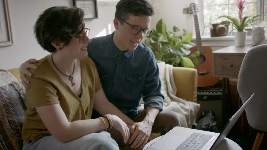 A couple sit on the couch smiling talking and waving to their friends on a video call on the laptop