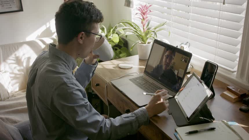 A man sitting video chatting on a laptop with a stylus in his hand using a tablet