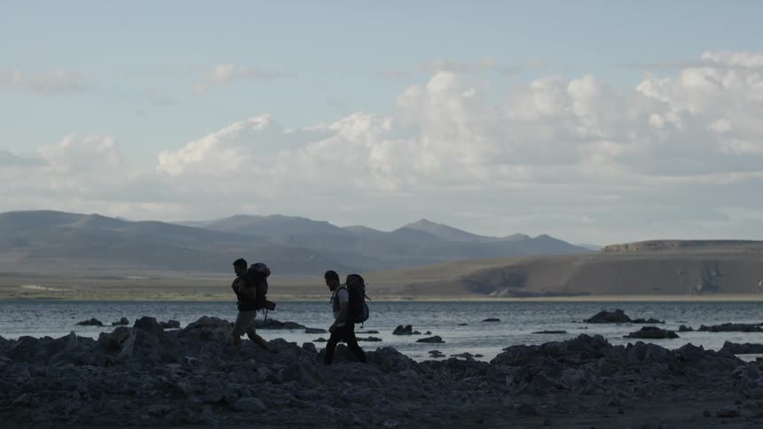 Hikers with backpacks walk over rocks beside the water