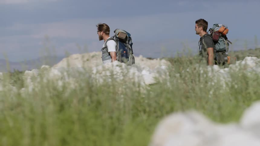 Two men wearing backpacks hike around rocks on a sunny day