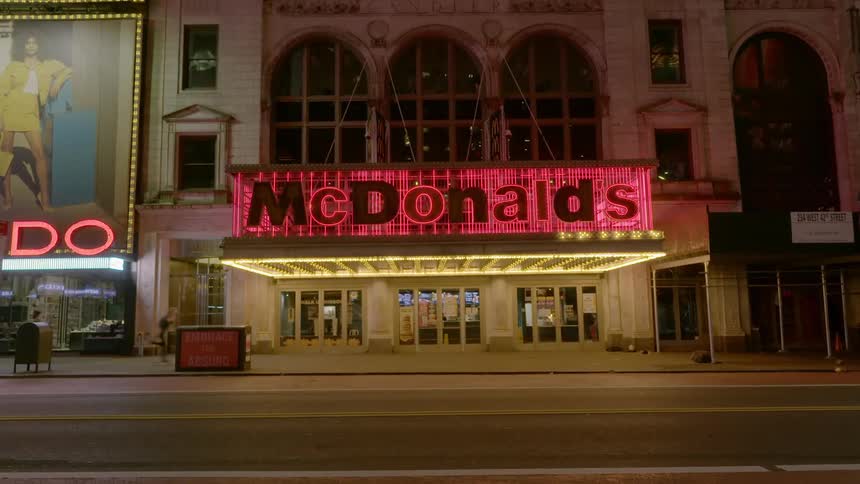 Skateboarder and pedestrian cross in front of buildings with neon signs at night