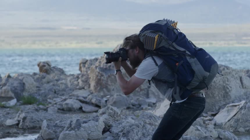A man wearing a backpack stands on a rocky shore taking photographs