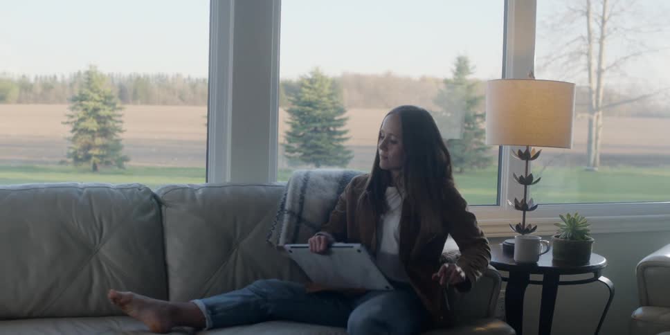 A woman walks into her living room carrying a laptop and mug and sits down to use it