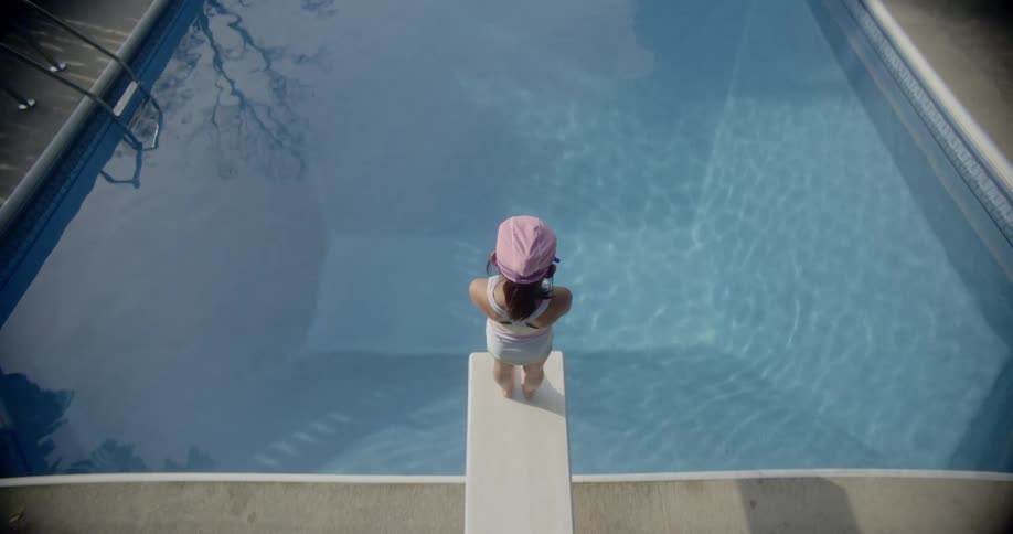Girl wearing swimsuit stands on diving board looking over swimming pool in house backyard