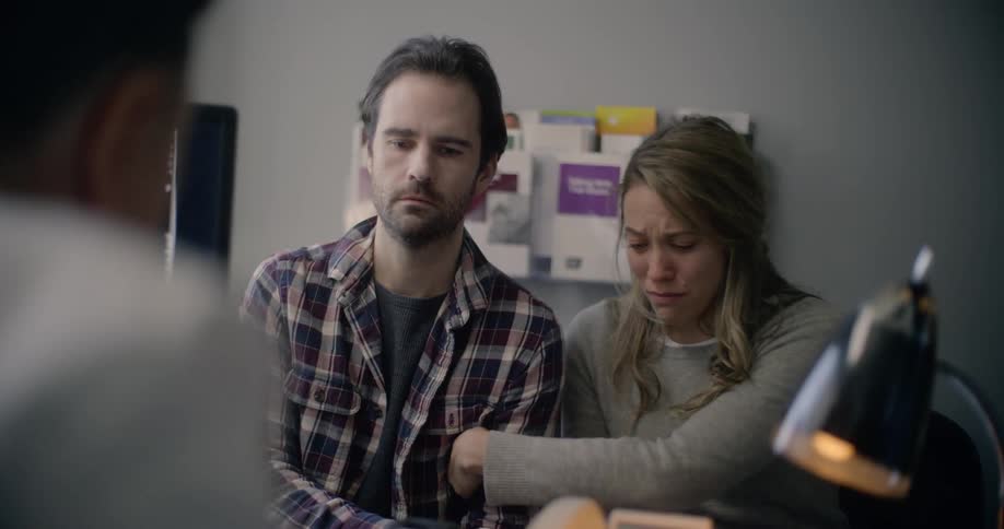 A crying woman leaning on the shoulder of a distraught man as they sit across from a doctor in a doctors office