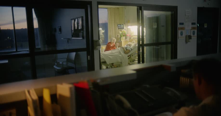 A nurse sits outside a hospital room where a woman holds her husbands hand as he lies in bed