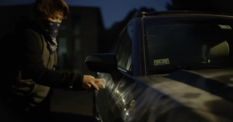 A teenage boy vandalizes a parked car by tagging it with spray paint at night
