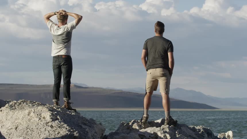 Two men stand on rocks and look out at the water on a cloudy day