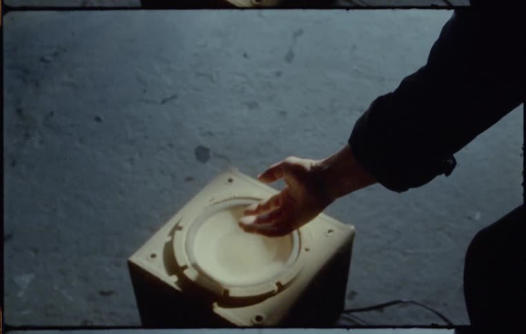Person reaches for cream colored speaker vibrating on concrete floor