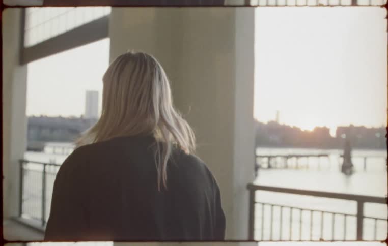 Woman walks along covered area next to water across from city with sun on the horizon