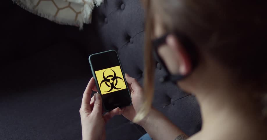 A woman sits on a couch looking at a large symbol on her cell phone and wearing a face mask