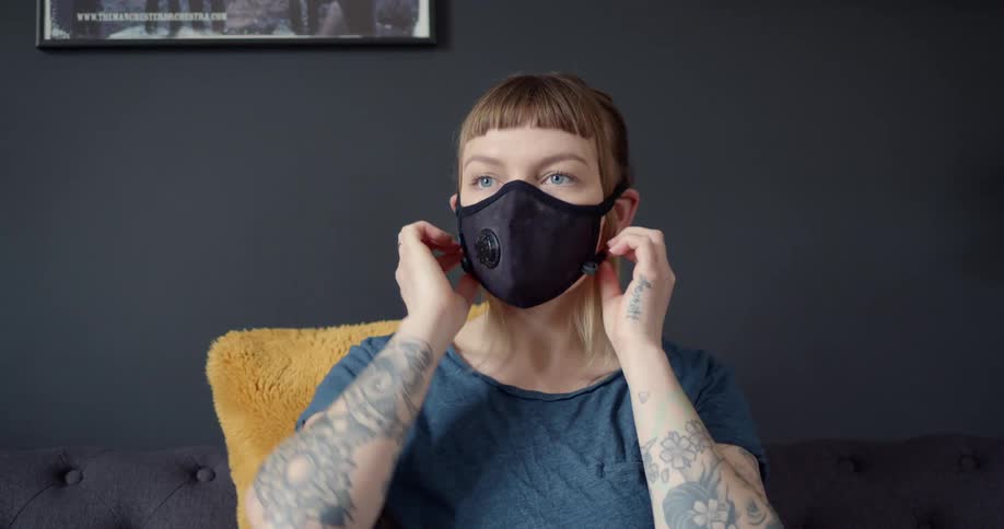 A woman with tattoos on her arms sitting in a chair puts a cloth filtered mask on her face