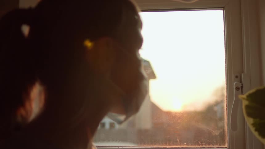 A woman in a medical face mask looking out of a small window at the sunset