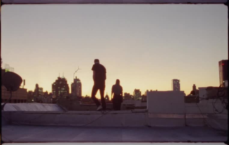 A man and woman on a rooftop hop over the barrier and look out at the city skyline at sunset