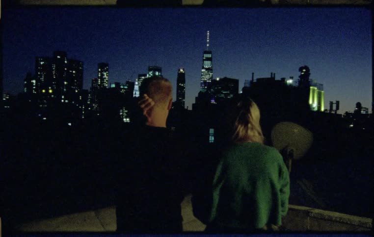 Man and woman standing on the rooftop of a building watch the city skyline at night