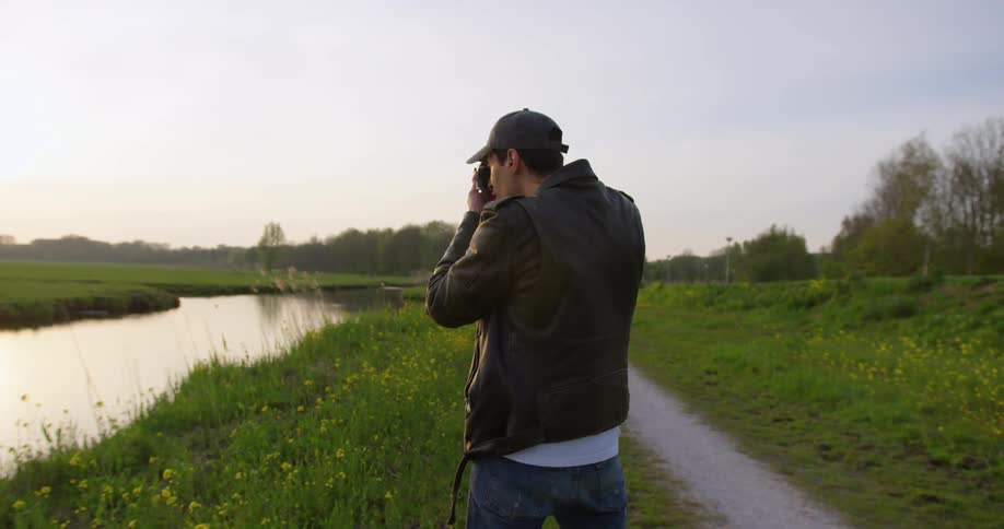 A man holds a camera to his face standing beside a rural pond
