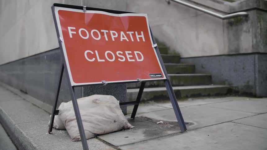 A sign is posted on a city sidewalk showing that the path is closed to pedestrians