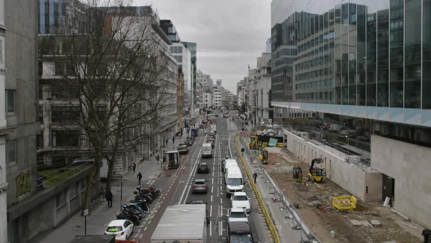 Cars and trucks travel on a city street with pedestrians on sidewalks and construction vehicles on one side