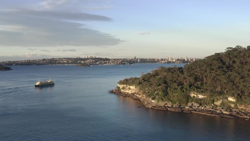 A large ferry moves through the water past a hilly shore heading towards a coastal city