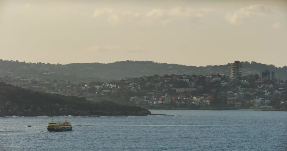 A motorboat passes a ferry traveling in the water off the coast of a city as waves splash up on the rocky shore