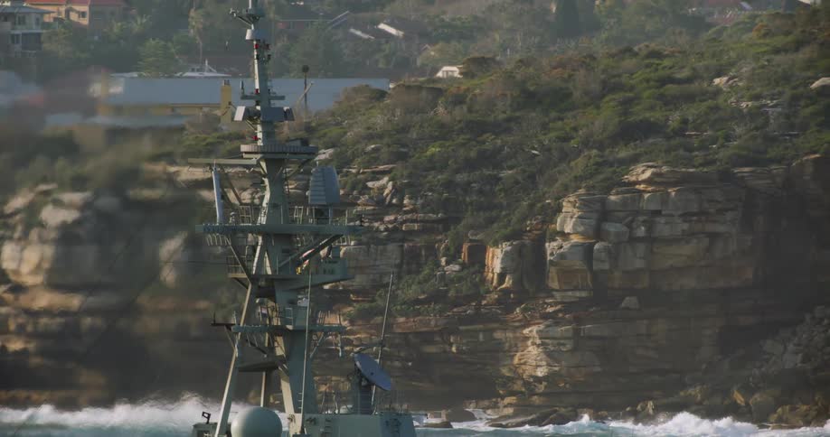 A ships antenna tower with satellite dishes as the ship passes cliff and ocean waves splashing on rock