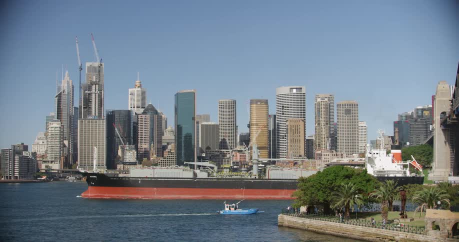 A cargo ship travels under a bridge and past a coastal city skyline on a blue sky day