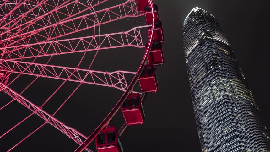 A ferris wheel with closed gondola cars turns with a skyscraper behind it at night