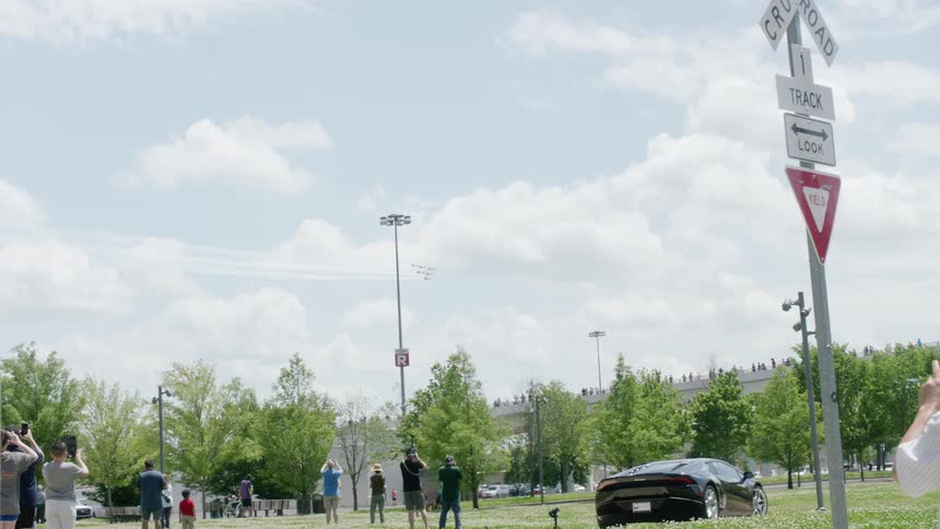 A crowd stands in a public park and watches and photographs military jets flying overhead in formation