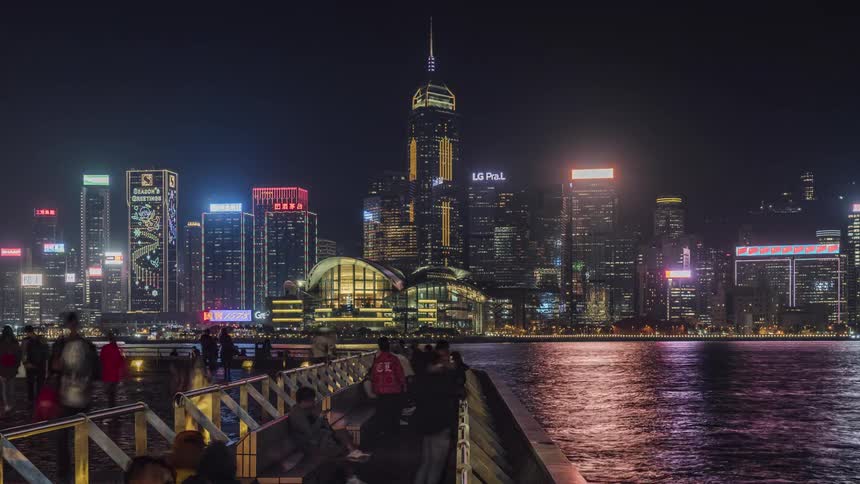 Tourists walk and sit on the waterfront looking at and photographing the colorfully lit hong kong skyline