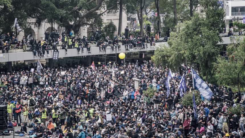 A large crowd gathers and waves flags at a rally in a public park of a city
