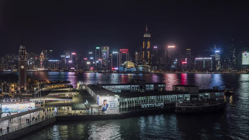 People walk along the waterfront and pier as the city skyline flashes with colorful lights