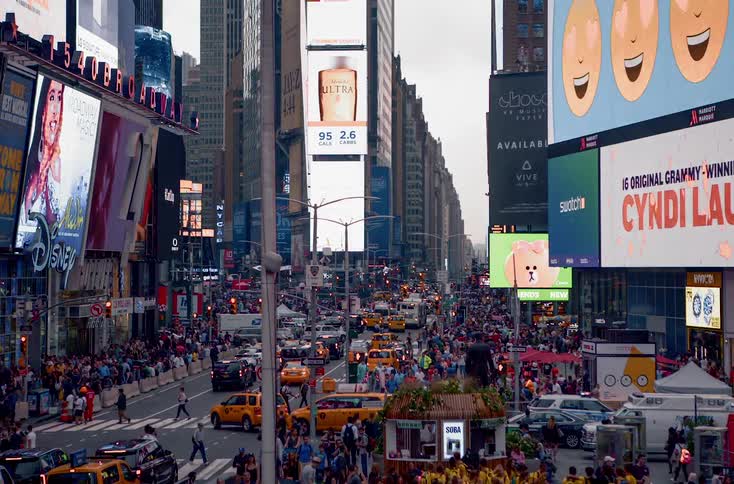 Images and text flash on electronic billboards above a crowded busy city street