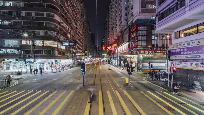 Cars and buses travel in a busy city at night with crowds of people walking and crossing streets