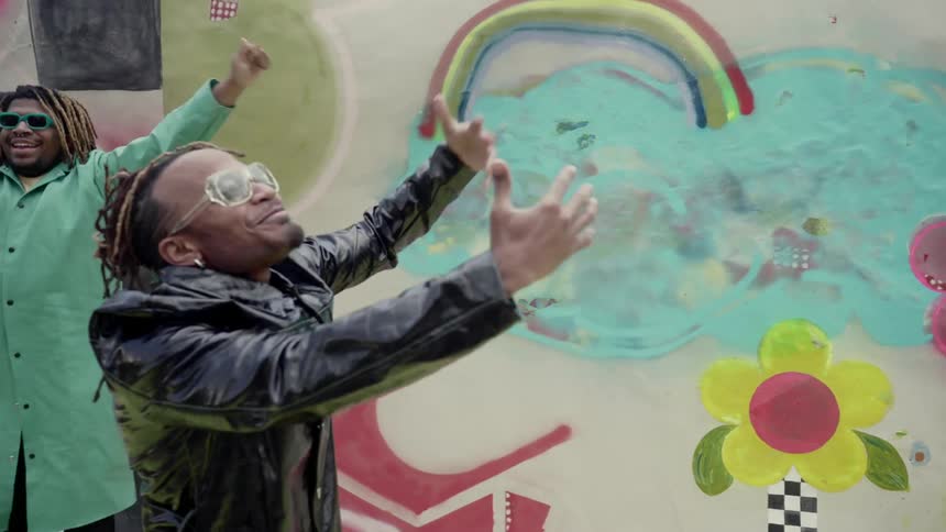 Two black men rap and dance in front of a wall with color artwork drawn on