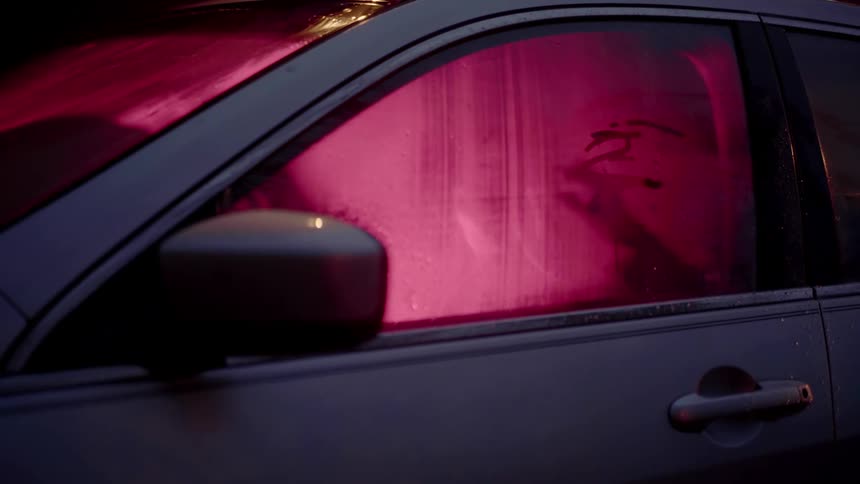 A person sitting in a car at night draws pictures and lines in the steam on the window