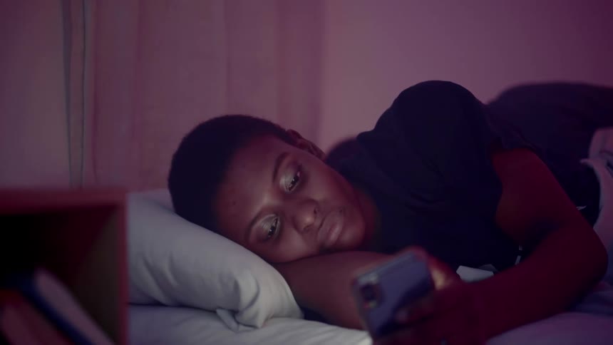 Black woman lying in the bed uses her smartphone in her room