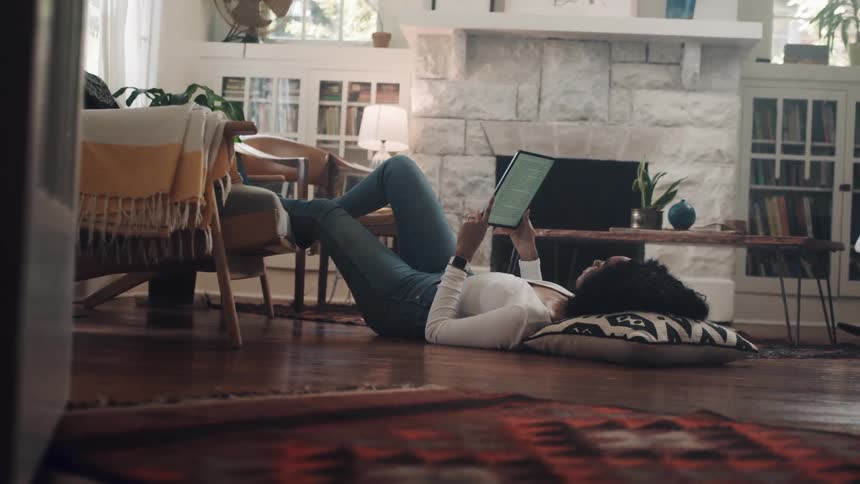 Black woman lying down on the floor uses a tablet in the living room at home